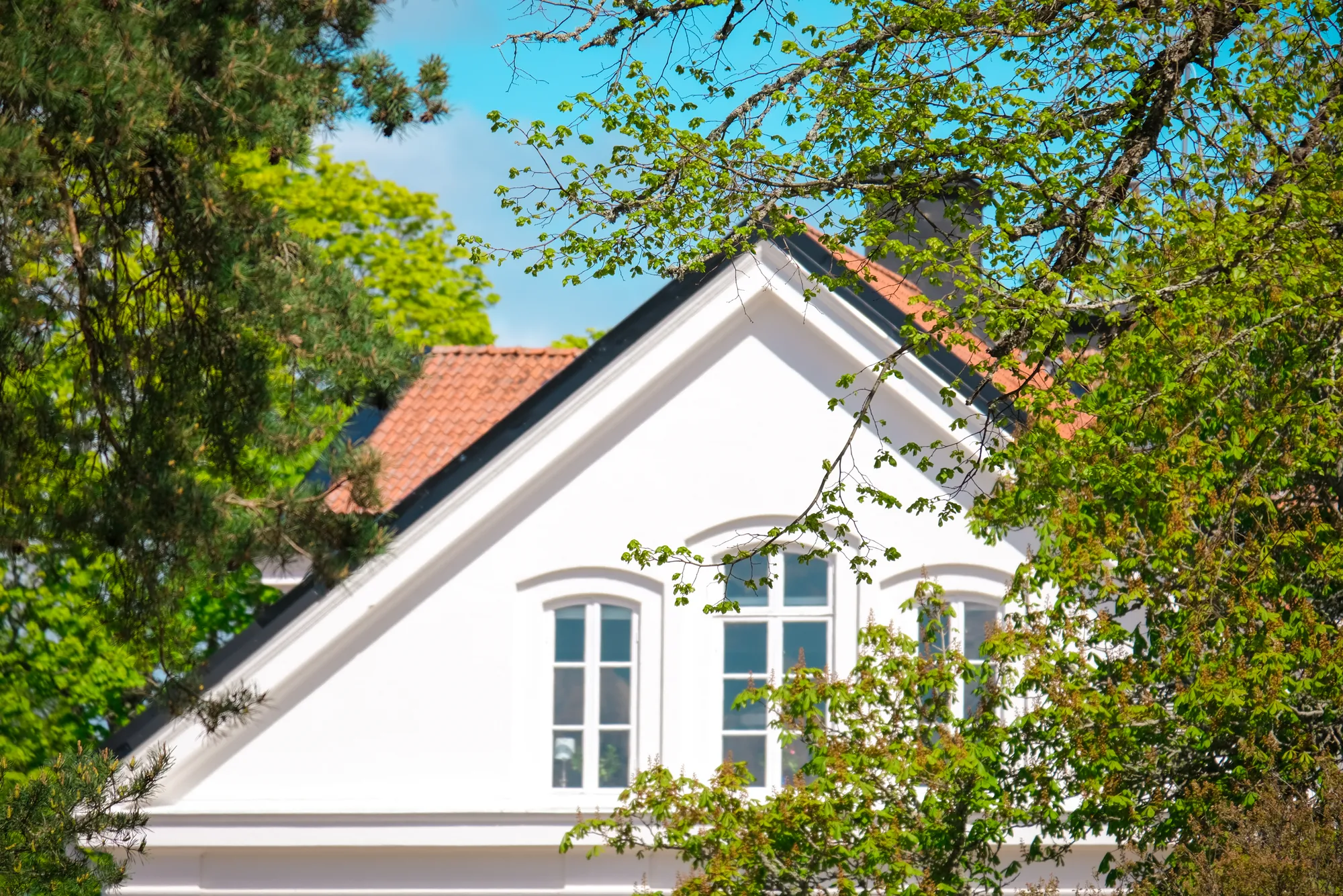 Den vita gaveln på ett hus med tegeltak och spröjsade fönster, omgivet av grönskande träd.