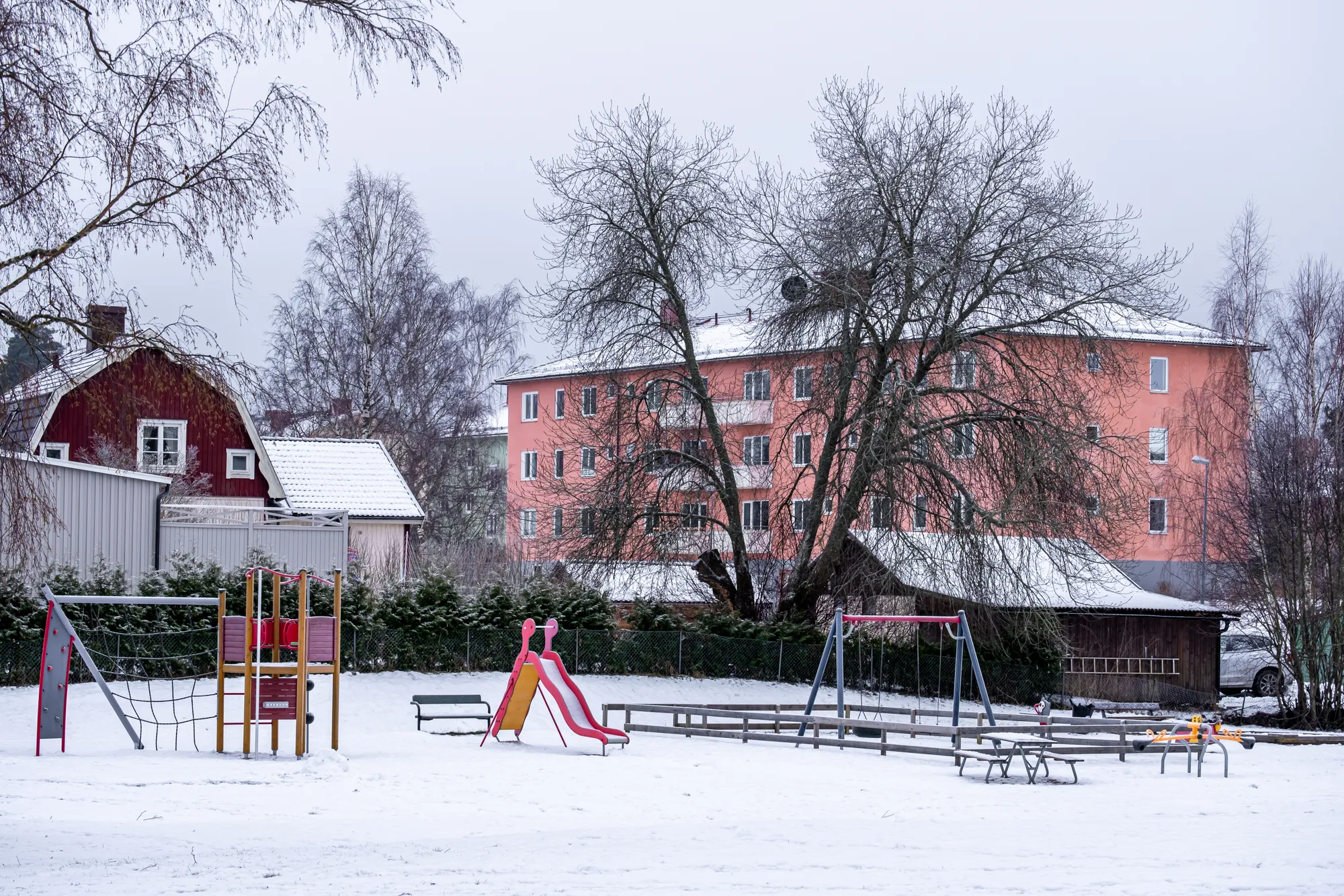 En snötäckt lekplats med rutschkana och gungor framför en rosa flerbostadshus och en röd lada.